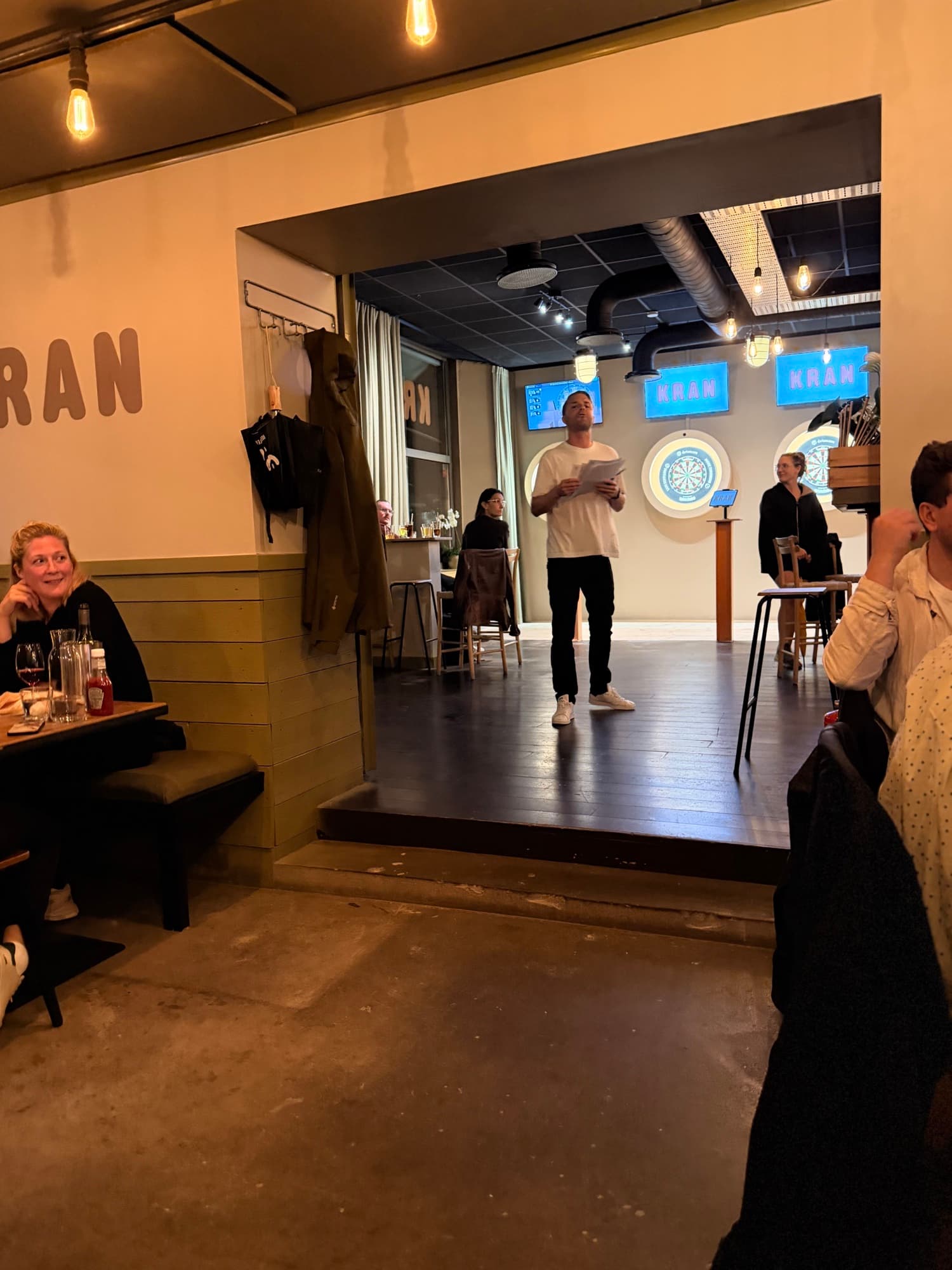Quizvärden står med frågorna i handen framför dartrummet och KRAN-skyltarna